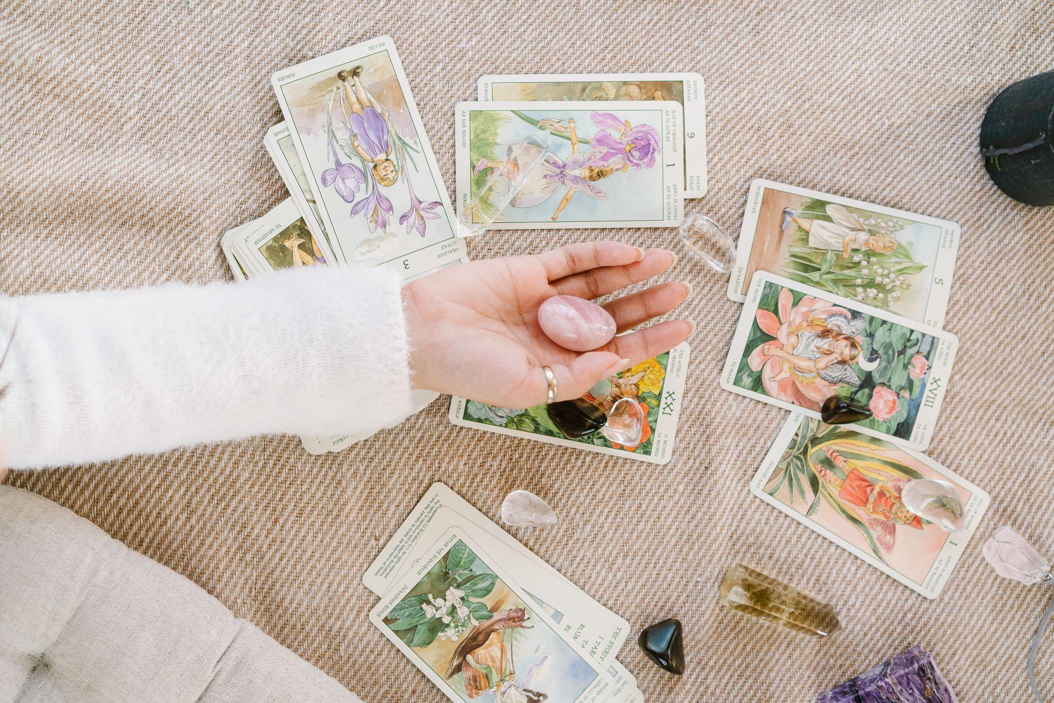a person’s outstretched hand holding a crystal while an assortment of tarot cards and crystals lie on the surface below it